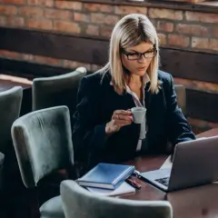 Kobieta w okularach pracuje na laptopie, popijając kawę. Na stole leży notatnik, być może planuje swój **konto Aurum**.