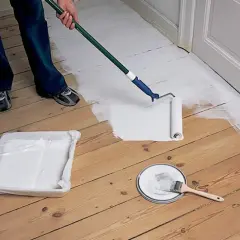 Person målar ett trägolv vitt med en roller. En färgtråg och en burk med färg står bredvid.