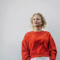 Sigrid Johnson, med sitt blonda, lockiga hår, bär en orange tröja och tittar drömmande bort.
