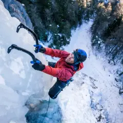 Uśmiechnięty mężczyzna w czerwonej kurtce i niebieskim kasku wspina się po lodospadzie, używając czekana. To ekscytująca wspinaczka lodowa.