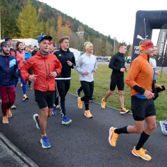 Grupa biegaczy w kolorowych strojach, jak z filmu o bieganiu, startuje na trasę. W tle las i bramka mety Puma.