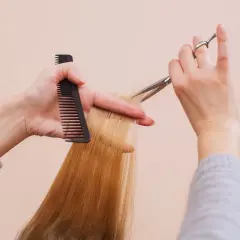 Hände mit Kamm und Schere schneiden blonde Haare. So kannst du kaputte Spitzen selber schneiden und deinem Haar neuen Glanz verleihen.