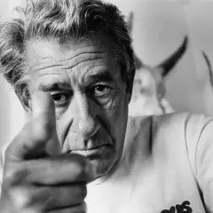 Helmut Newton, z palcem wskazującym w obiektyw, patrzy intensywnie. W tle widać fragmenty jego charakterystycznych prac.