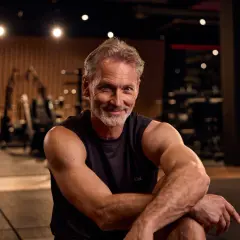 Ein lächelnder Mann mit grauem Haar und Bart sitzt im Fitnessstudio. Er zeigt, dass Muskelaufbau ab 50 möglich ist.