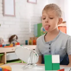 Chłopiec z wysuniętym językiem patrzy w lustro, obok kolorowe klocki.