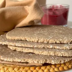 Una pila de pan sin levadura, rústico y con trozos de avena, junto a un vaso de mermelada de frutos rojos y una tela marrón.