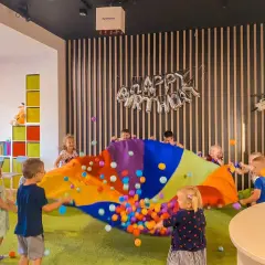Radosne urodziny dla dziecka w domu! Dzieci bawią się kolorowym spadochronem z piłeczkami, a w tle balony z napisem "Happy Birthday".