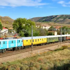 Un tren de Alsa, con vagones de colores, viaja por un paisaje montañoso. Este tren zaragoza logroño atraviesa un pueblo con casas blancas y tejados rojos.