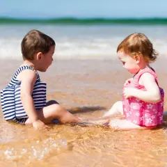 Dwoje dzieci w kostiumach kąpielowych z kamizelkami pływającymi siedzi na plaży.
