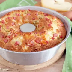 Gâteau salé végétarien doré, parsemé de dés de jambon et de fromage, dans un moule à savarin. Des œufs et du fromage sont visibles en arrière-plan.