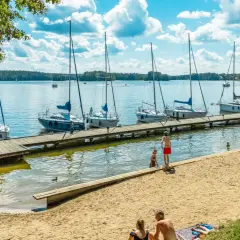 Letnie popołudnie na **jeziorach na Mazurach**. Drewniane molo, zacumowane żaglówki i plażowicze cieszący się słońcem.