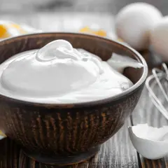 Un bol rempli de crème fouettée, blanc en neige, prêt à être utilisé pour une pâtisserie. Des œufs et un fouet sont visibles à proximité.