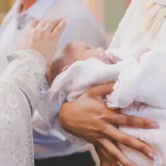 Dłoń trzyma dziecko owinięte w biały ręcznik podczas ceremonii chrztu.
