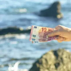 Dłoń trzyma banknoty euro na tle morza. W Czarnogórze obowiązuje euro, więc te pieniądze przydadzą się na wakacjach.