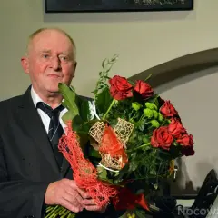 Starszy mężczyzna w garniturze trzyma bukiet czerwonych róż i zielonych kwiatów.