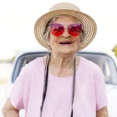Une femme souriante, chapeau de paille et lunettes roses, prête pour partir seule en vacances à 60 ans. Une voiture ancienne en arrière-plan.