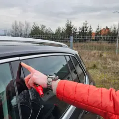 Mężczyzna w czerwonej kurtce wskazuje palcem na drzwi swojego pierwszego samochodu, trzymając w dłoni czerwony przyrząd.