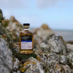 Bouteille de whisky "Elements of Islay" posée sur des rochers moussus, évoquant l'atmosphère sauvage de la distillerie Islay.