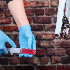 Hands in blue gloves bleed bike brakes, using a syringe filled with red fluid. This shows how much brake fluid to bleed brakes for optimal performance.