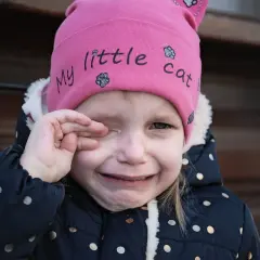 Mała dziewczynka w różowej czapce z napisem "my little cat" płacze, pocierając oko. Wygląda, jakby coś jej wpadło, może opiłek w oku.