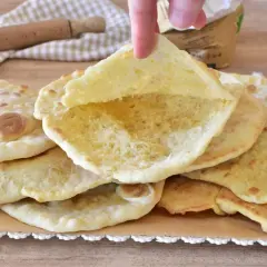 Un dito solleva un soffice pane in padella, mostrando la sua interna alveolata. Altri dischi dorati riposano su carta forno.