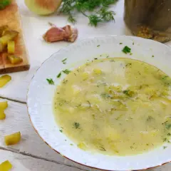 Gotowy przepis na zupę ogórkową. Na desce pokrojone ogórki, obok talerz z gotową zupą, słoik z ogórkami i koperek.