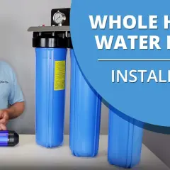 Man in a cap and polo shirt stands next to a whole house water filter system, ready for installation.
