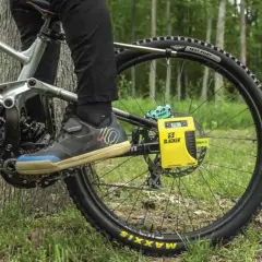 A person uses a yellow Slacker digital scale for rear shock measurements on a mountain bike, with a forest background.