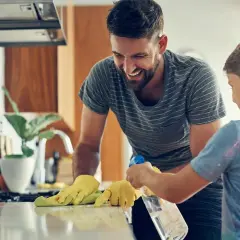 Padre e hijo ríen mientras aprenden como limpiar tu casa. El niño rocía la encimera mientras el padre la limpia con un paño.