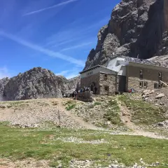 Schronisko pod masywnym **Naranjo de Bulnes**. Grupa turystów odpoczywa na tle skalistych szczytów i błękitnego nieba.
