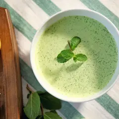 Un bol de sauce à la menthe fraîchement préparée, garni de feuilles de menthe. À côté, une tranche de citron et une gousse d'ail sur une planche en bois.