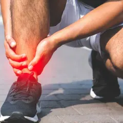 Osoba trzymająca się za bolesną, czerwoną kostkę w bucie sportowym.