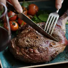 Ein saftiges Lamm-Steak wird mit Messer und Gabel angeschnitten. Dazu passen gerösteter Knoblauch, Kirschtomaten und ein Glas Rotwein. Welcher Wein zu Lamm? Ein kräftiger Rotwein!