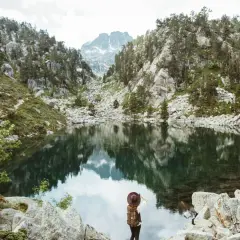 Una excursionista admira un lago sereno en el **parque nacional de los Pirineos**, rodeado de montañas rocosas y pinos.