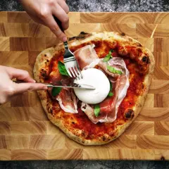 Une pizza burrata avec du prosciutto et du basilic, prête à être dégustée. Des mains coupent la burrata crémeuse.