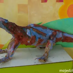 Kolorowa dinozaury praca plastyczna z papieru, z otwartą paszczą i ostrymi zębami, na tle malowanej ściany.