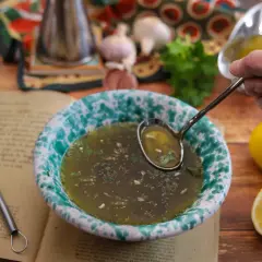 Un mestolo raccoglie il salmoriglio da una ciotola, pronto per la ricetta. Ingredienti freschi e un libro di cucina completano la scena.