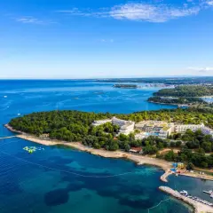 Hotel Delfin Plava Laguna z lotu ptaka, otoczony zielenią i błękitnym morzem. Widoczne baseny, plaża i przystań z łodziami.