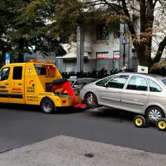 Żółty holownik odholowuje srebrny samochód. Kierowca dostał mandat za parkowanie.