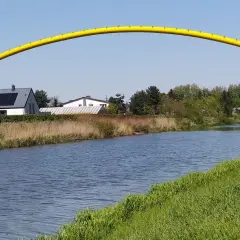 Żółty łuk mostu nad kanałem żerańskim, z domami i zielenią w tle.