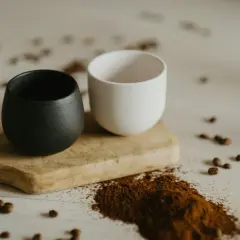 Dwa ceramiczne kubki, czarny i biały, na drewnianej desce. Obok rozsypana mielona kawa, która sugeruje, czy kawe sypana sie miesza.