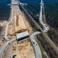 Budowa drogi ekspresowej S19 Via Carpatia w lesie. Widoczna nowa nawierzchnia, most, kolej i ruch samochodowy.