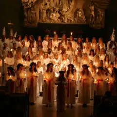 Stor grupp barn i vita dräkter och ljuskransar deltar i ett lucia firande i Stockholm.