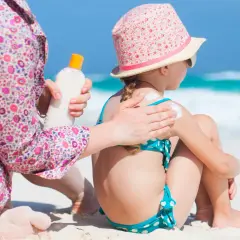 Mamma applica la crema solare a una bambina sulla spiaggia, prevenendo la dermatite da sabbia.