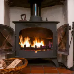Ein Holzofen mit brennendem Feuer. Daneben ein Korb mit **Holz** und ein Werkzeugset. Die **Asche** glüht im Feuer.
