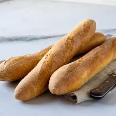 Tres baguettes doradas recién horneadas, listas para aprender como hacer baguette. Un cuchillo de madera descansa a su lado.