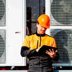 Technik w pomarańczowym kasku sprawdza dane na tablecie przy jednostkach zewnętrznych pompy ciepła, planując dofinansowanie.