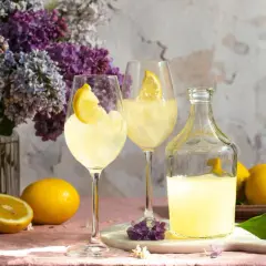 Erfrischendes Lemon Spritz Rezept: Zwei Gläser mit Eis und Zitronenscheiben, daneben eine Karaffe mit Zitronenlikör und frische Zitronen. Ein Blumenstrauß aus Flieder im Hintergrund.