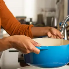 Une personne remplit une cocotte bleue sous un robinet pour faire bouillir de l'eau chaude.