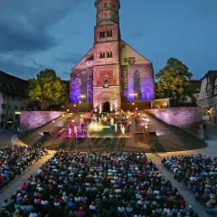 Bühnenaufführung vor historischer Kulisse in Hohenlohe. Ein großes Publikum verfolgt gespannt die Darbietung auf illuminierten Treppen vor einer Kirche.